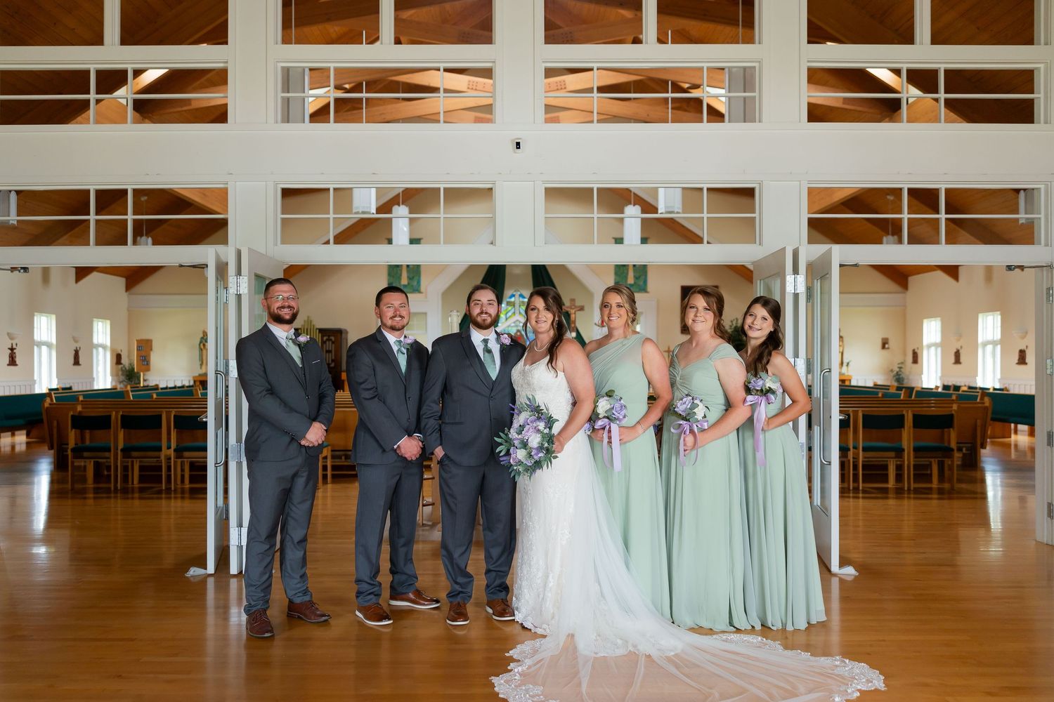 Wedding party poses in elegant venue, bride in white gown with bridesmaids in sage green dresses and groomsmen in gray suits.
