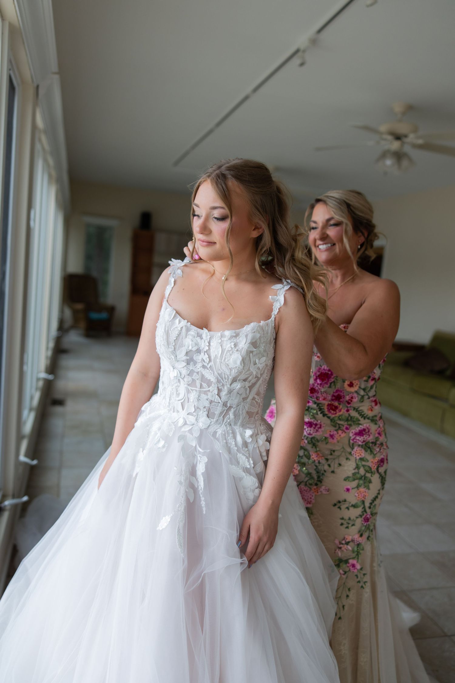Bride in white lace ball gown being helped by bridesmaid in floral dress during wedding preparation.