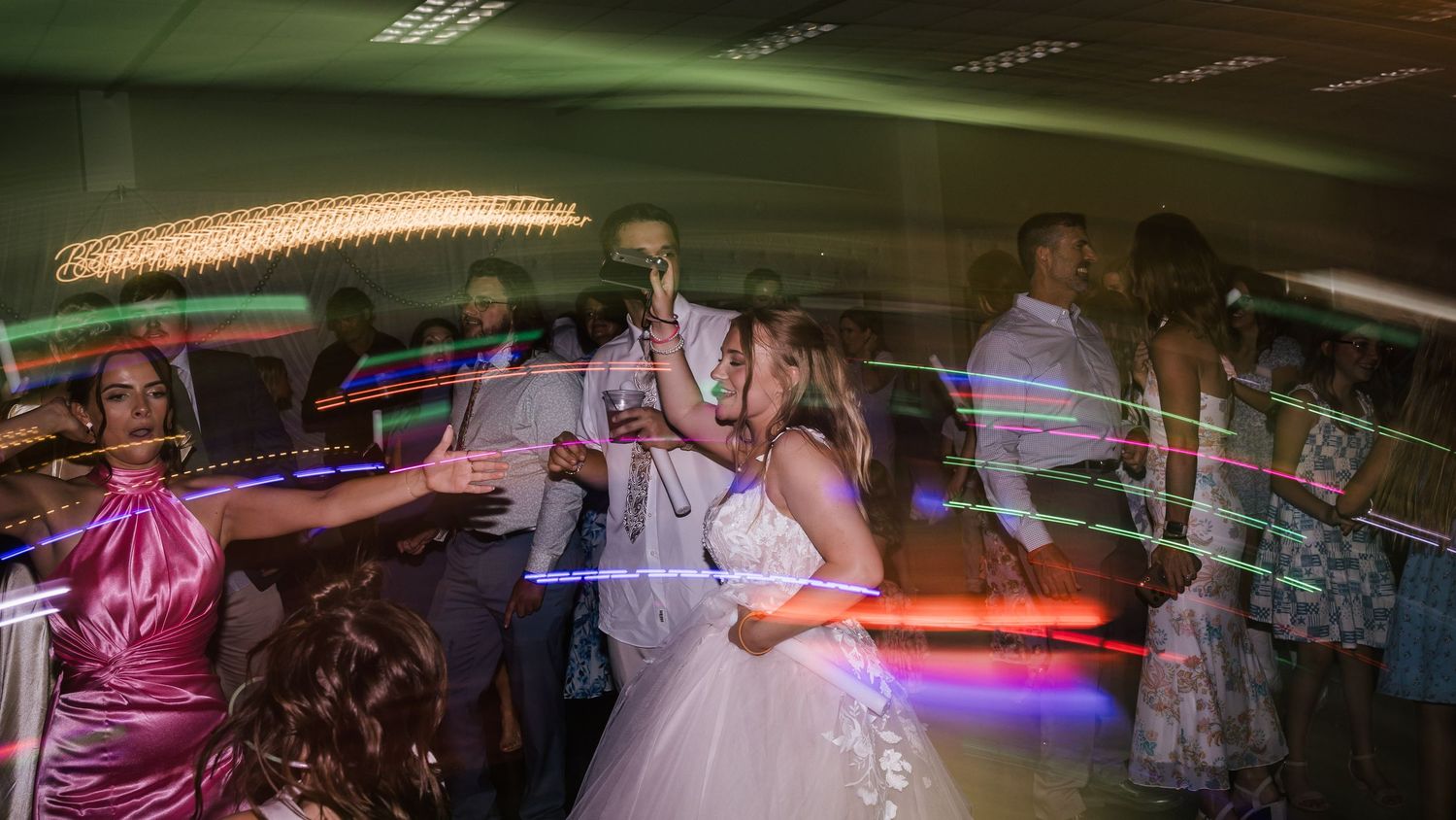Bride and groom share first dance surrounded by guests with colorful light streaks at reception.