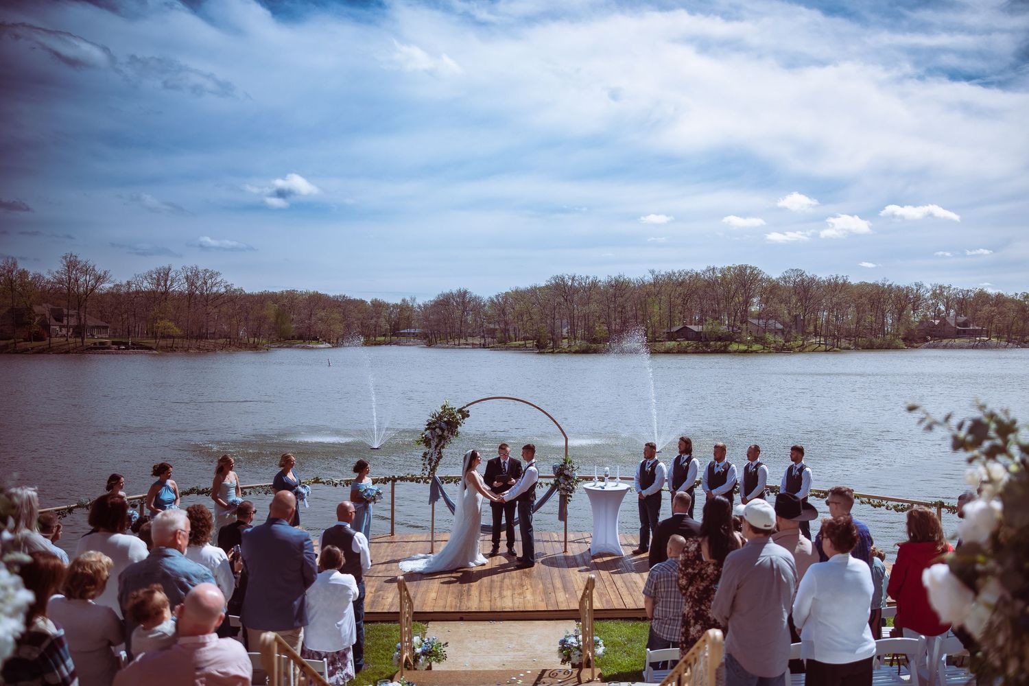 Outdoor lakeside wedding ceremony on dock with circular arch, wedding party, and guests under blue sky.