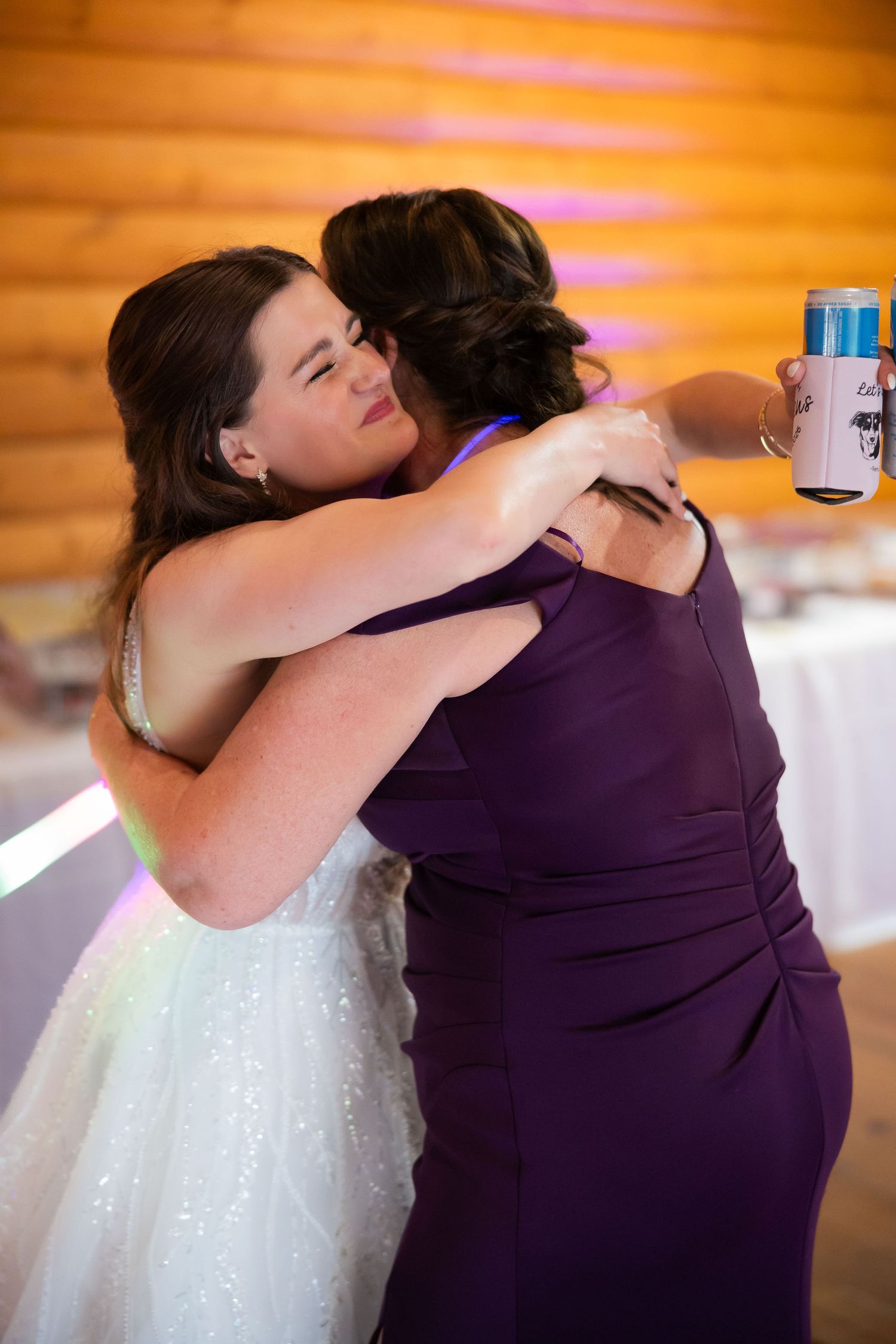 Bride in white sparkle gown shares heartfelt hug with bridesmaid in purple dress at wedding reception.