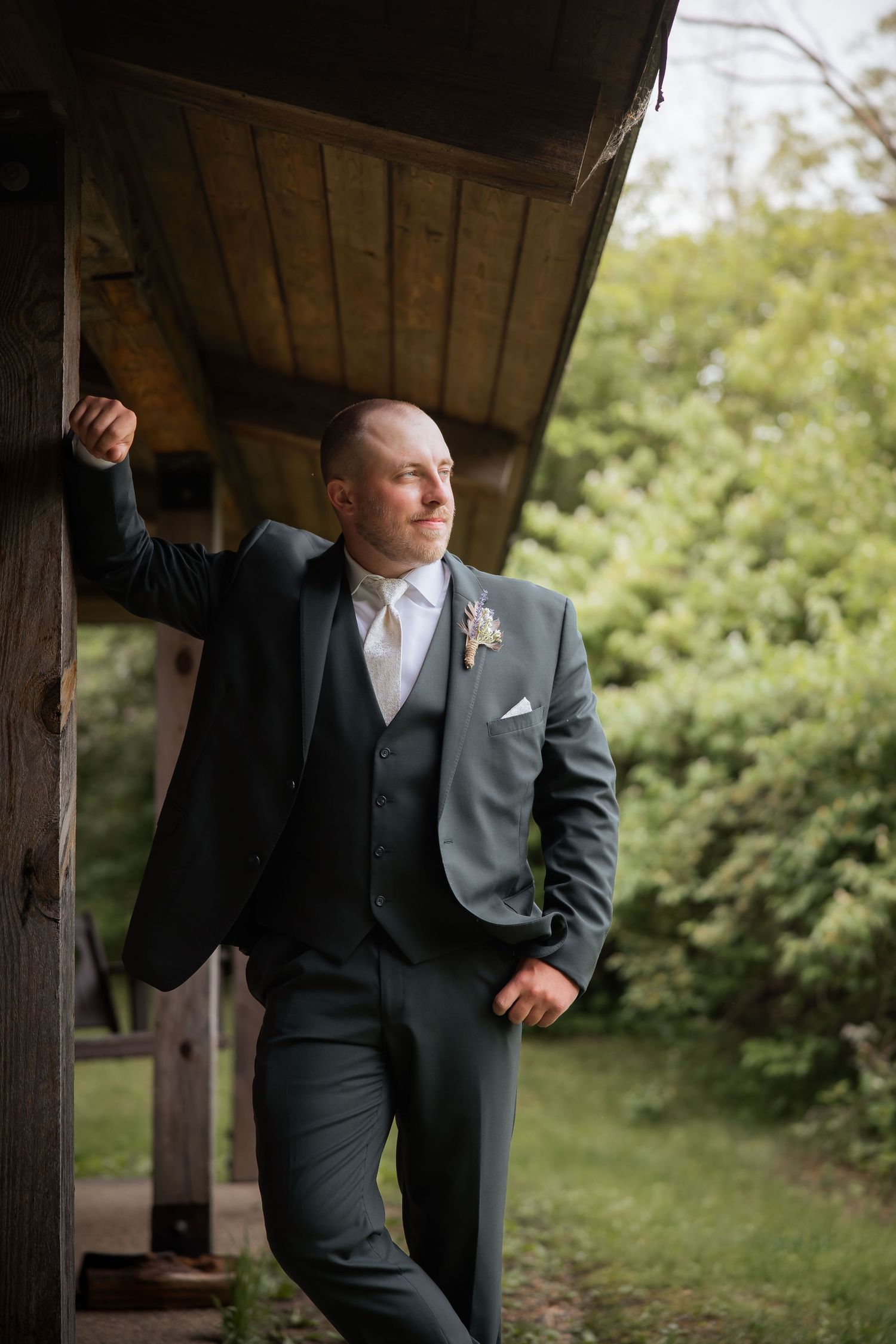 Elegant groom in gray three-piece suit with boutonniere leans against rustic wooden structure outdoors.