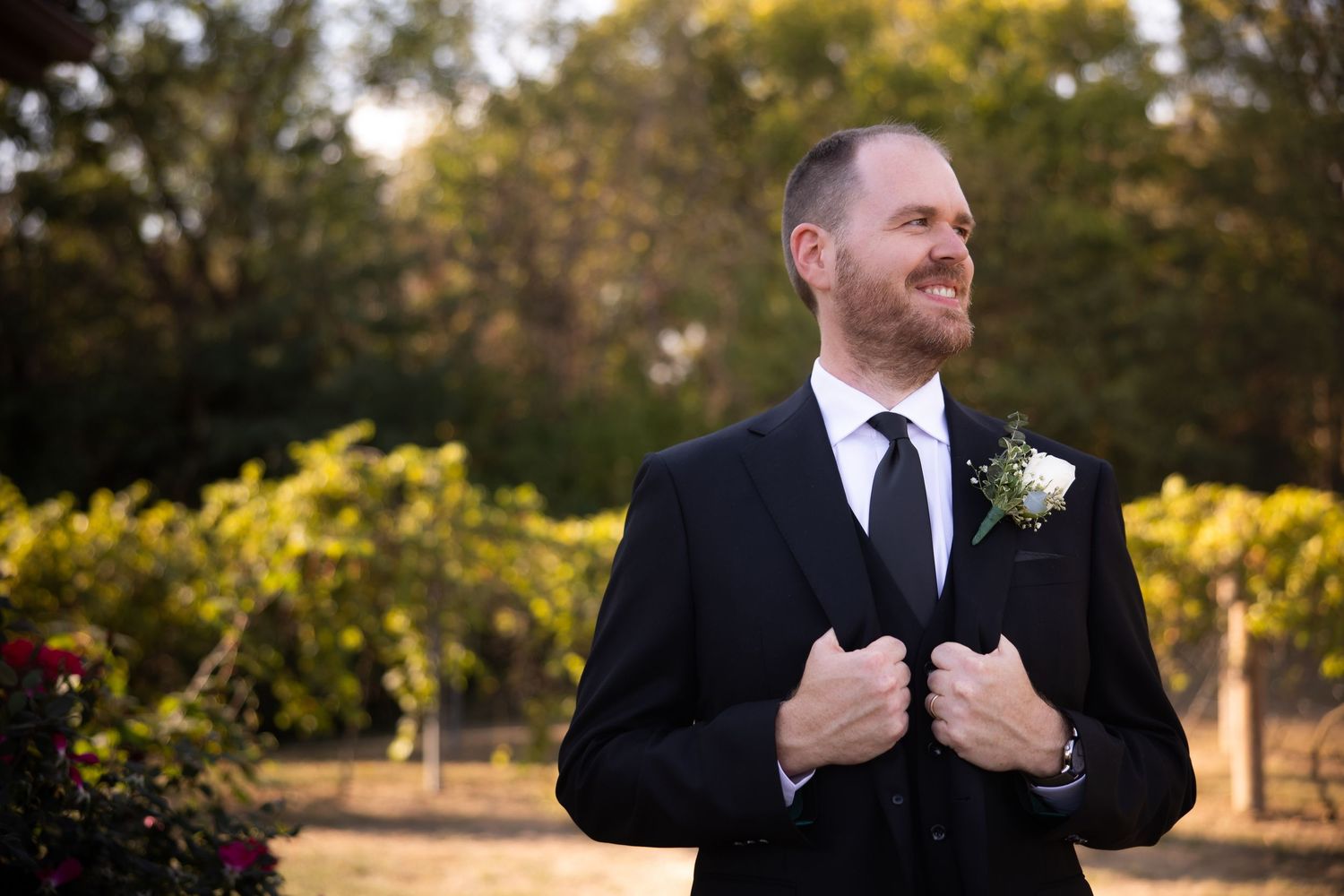 A person in formal attire smiles while adjusting their collar outdoors.
