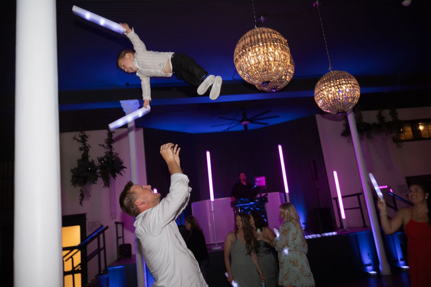 Wedding guests toss someone in the air under glittering disco balls and purple lighting.