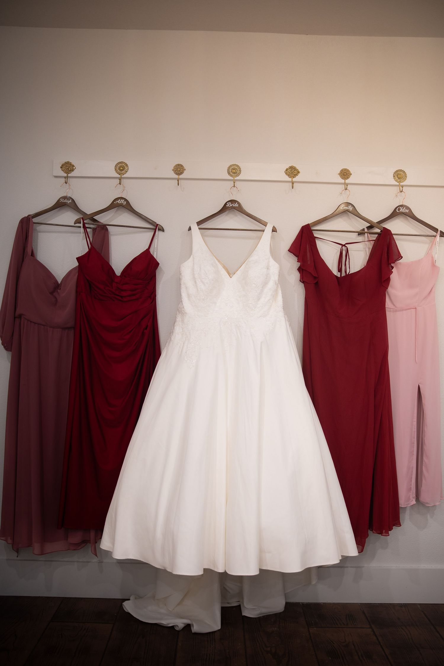 Wedding party dresses in burgundy and white hanging on a wall-mounted rack against a neutral background.