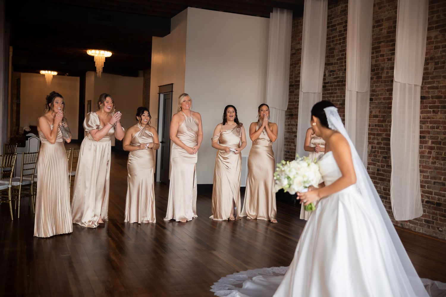A bridal party in champagne-colored dresses reacts with joy during a first look reveal moment at an indoor venue.
