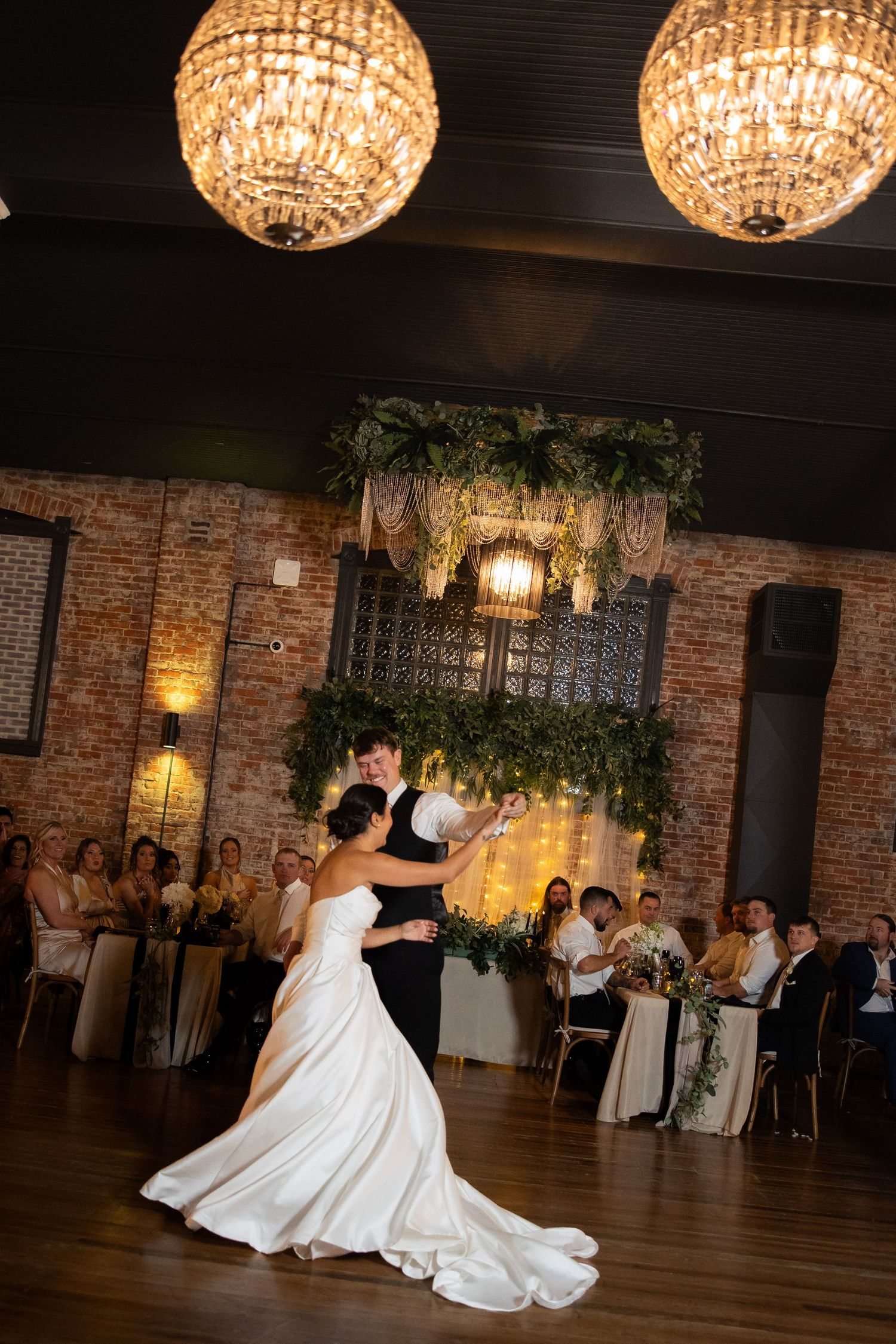 A romantic first dance moment captured under crystal chandeliers in an elegant brick-walled reception venue.