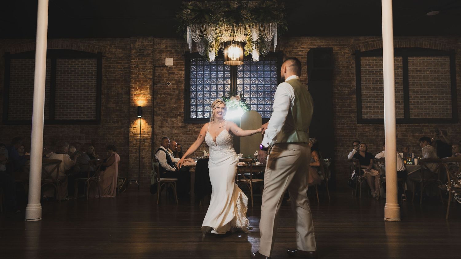 A couple shares their first dance in an elegant ballroom with warm lighting and stone archways.