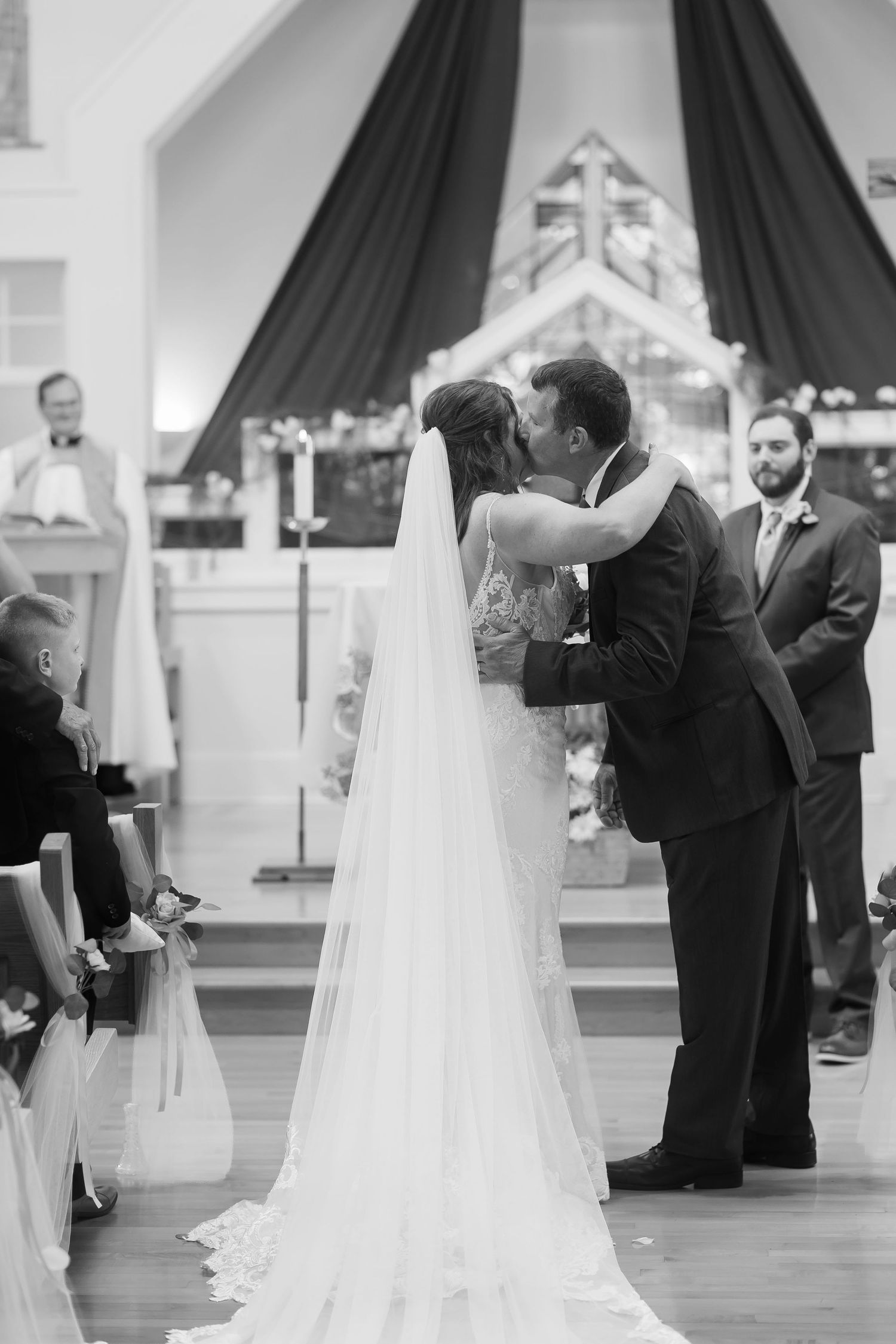 Black and white photo of bride and groom sharing first kiss at church altar during wedding ceremony.