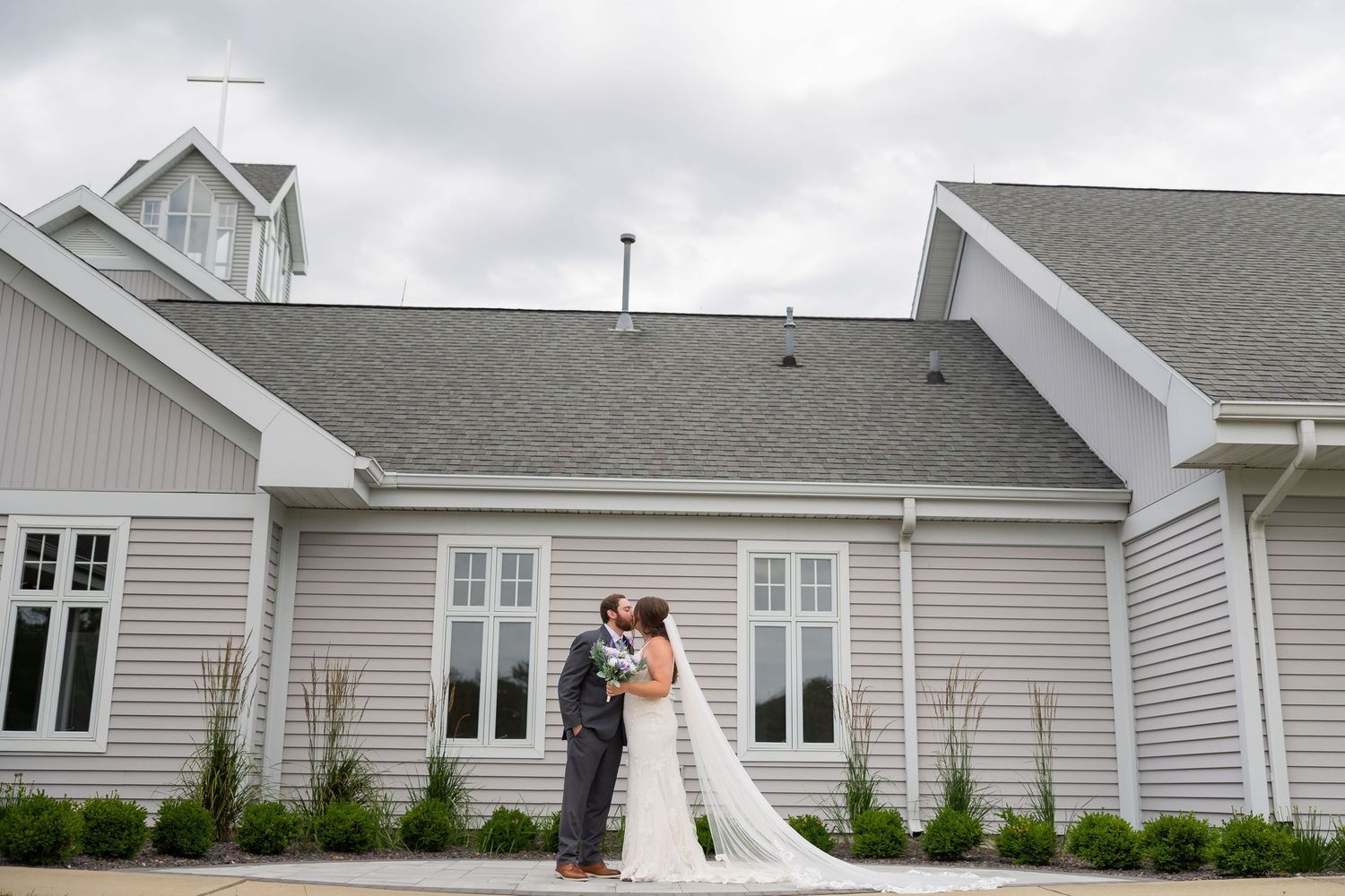 Bride and groom share a kiss outside a gray wedding venue on a cloudy day.