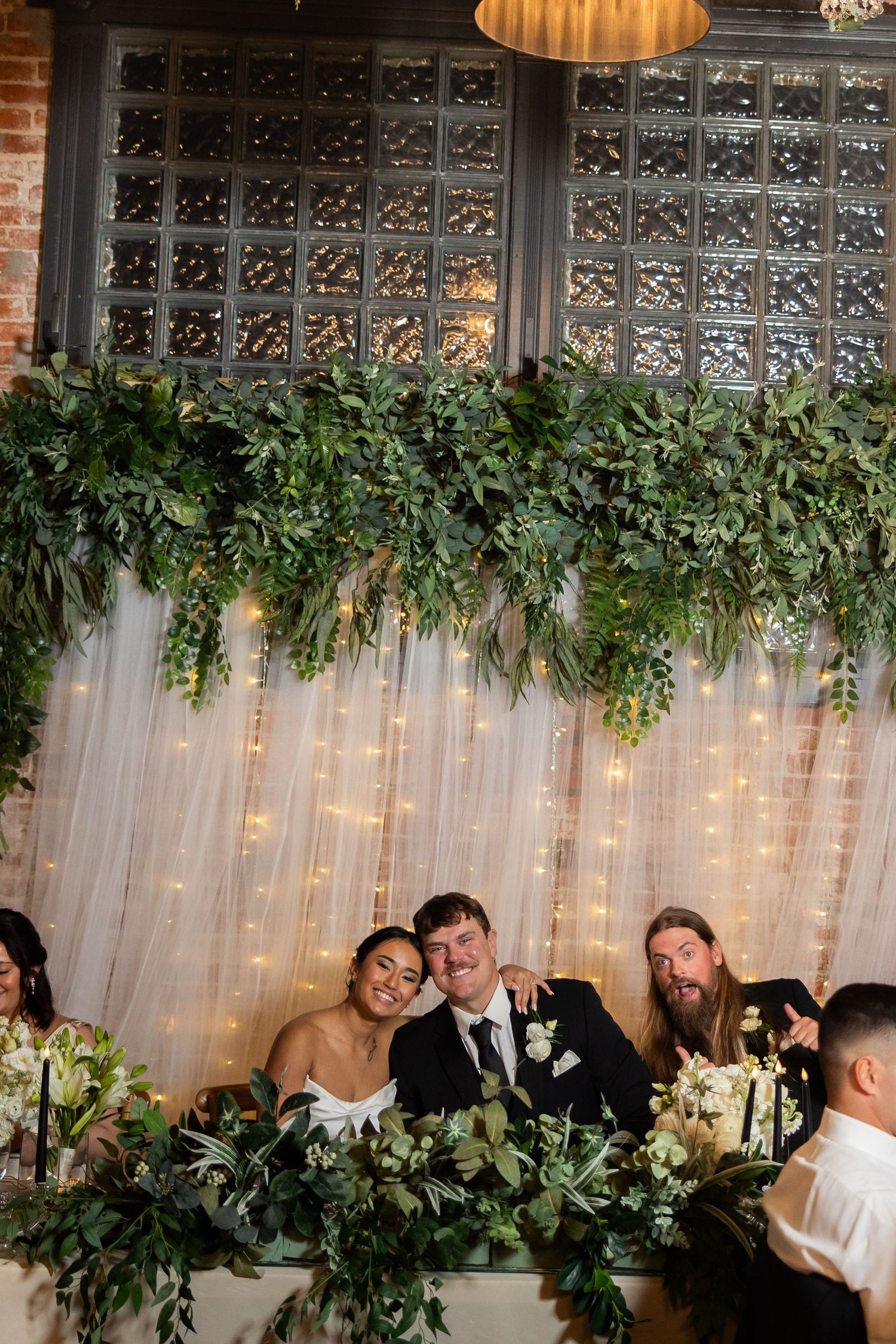 Wedding reception head table decorated with greenery, fairy lights and ornate metalwork backdrop.
