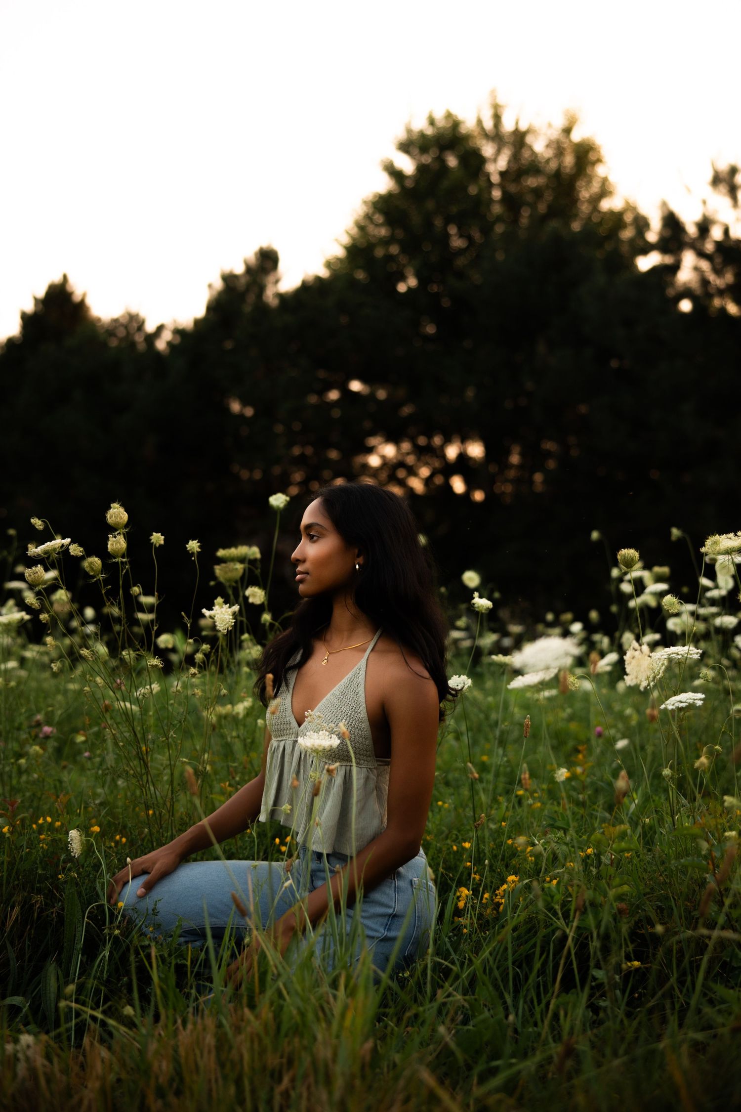 A young woman sits peacefully in a wildflower meadow at dusk, wearing a light camisole and jeans.