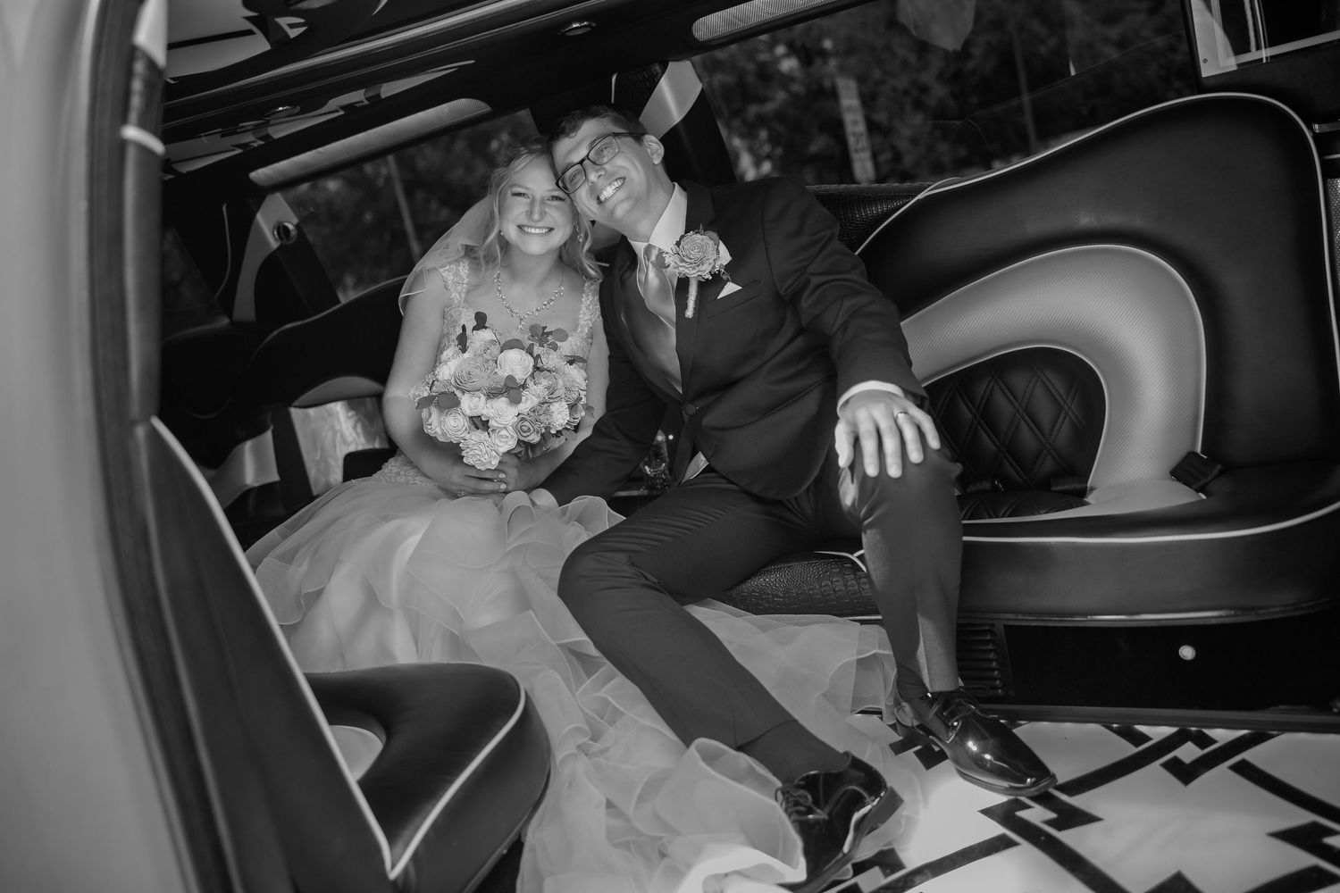 Black and white photo of a smiling bride and groom sitting together in a luxury limousine on their wedding day.