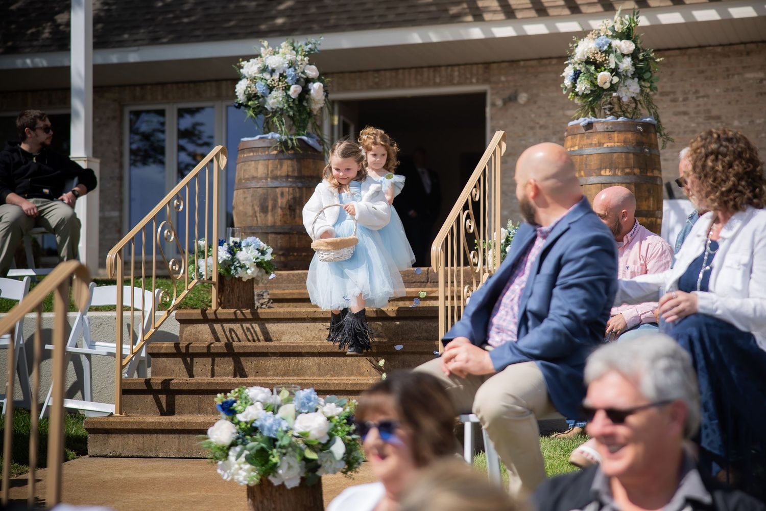 Two flower girls in white dresses descend outdoor wedding ceremony steps adorned with floral arrangements and barrel decorations.