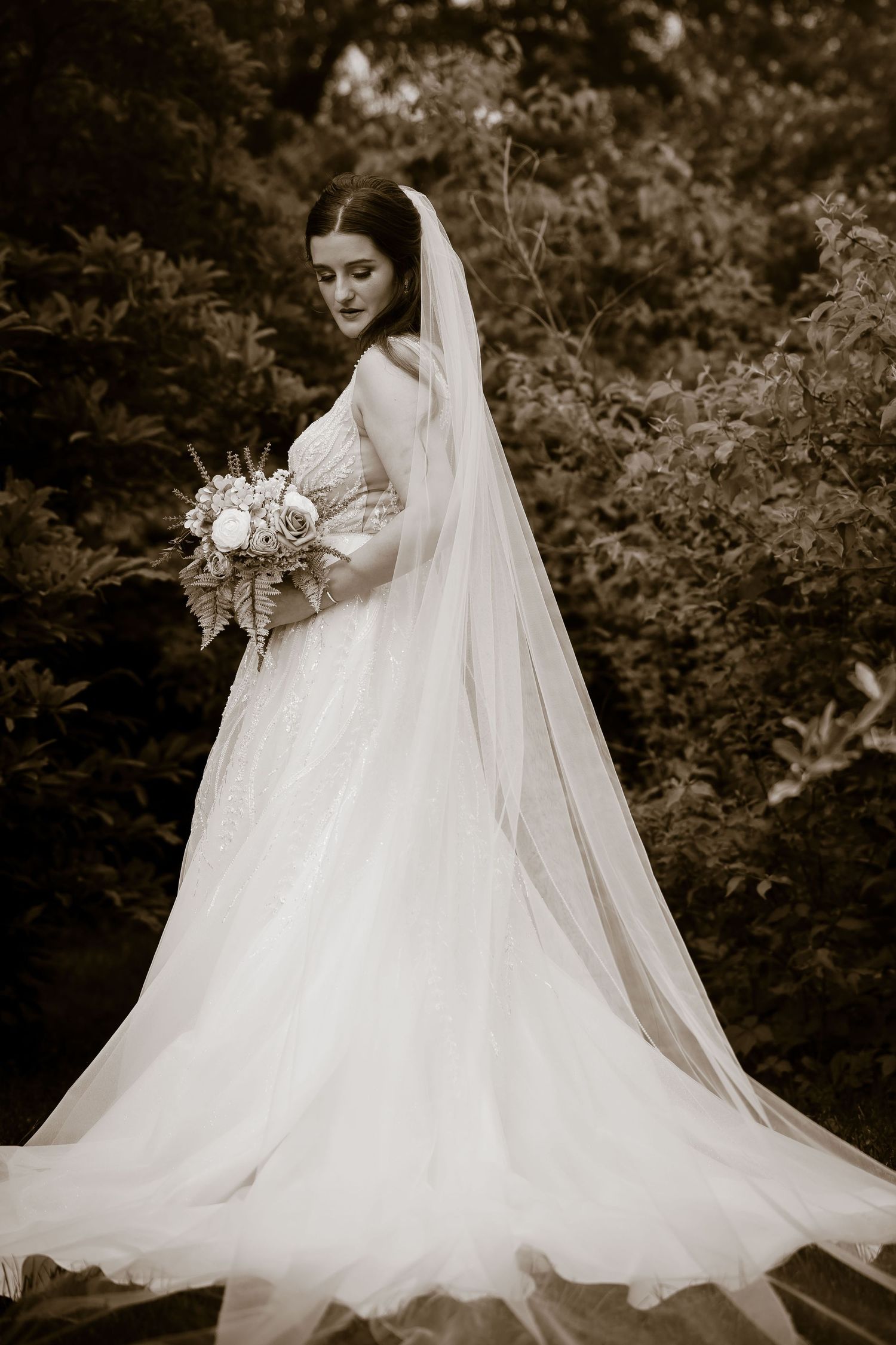 Elegant bride in a flowing white gown and long veil holds a bouquet in a sepia-toned outdoor portrait.