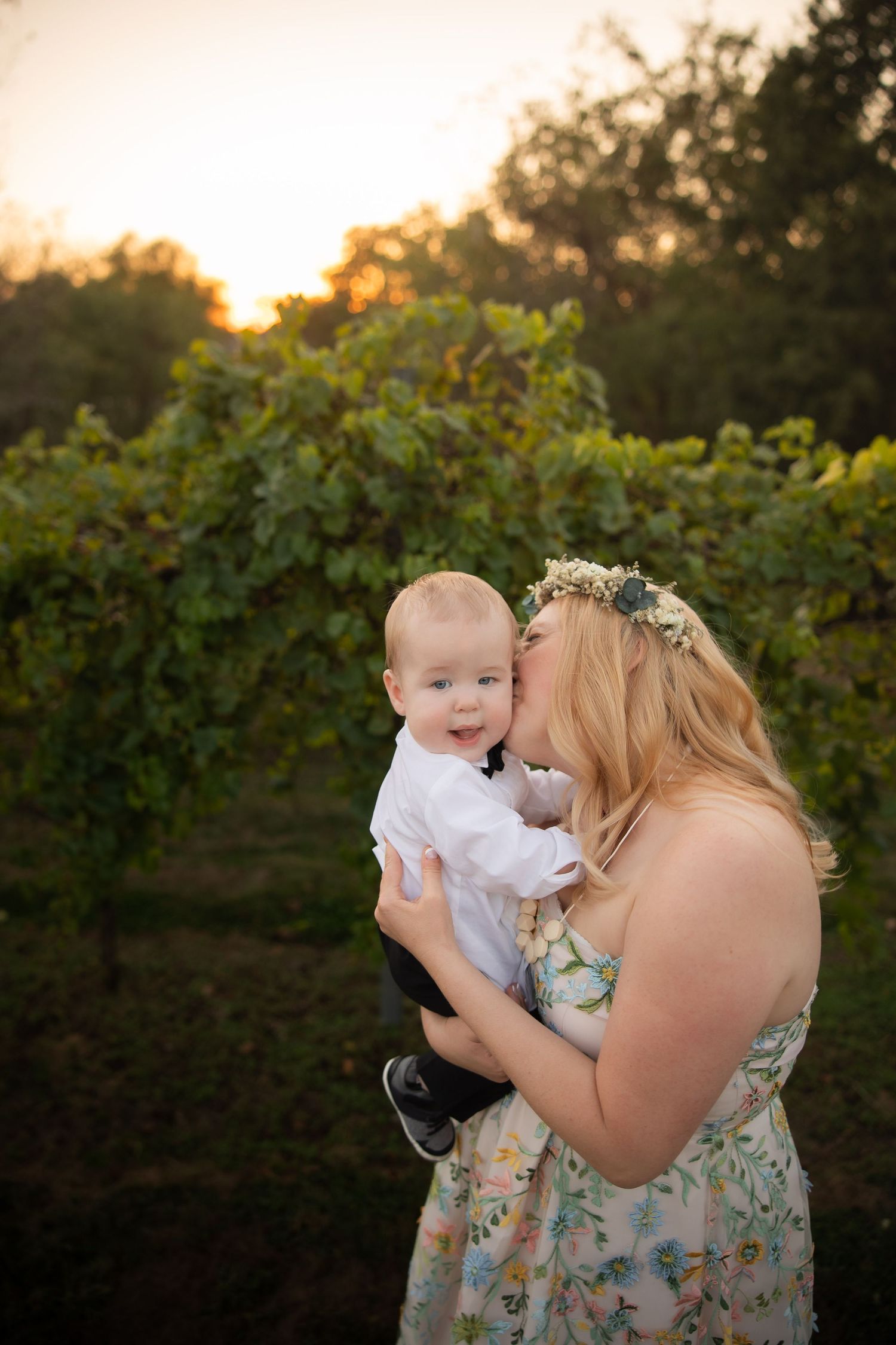 Mother and baby share a tender moment in front of vineyard at sunset.