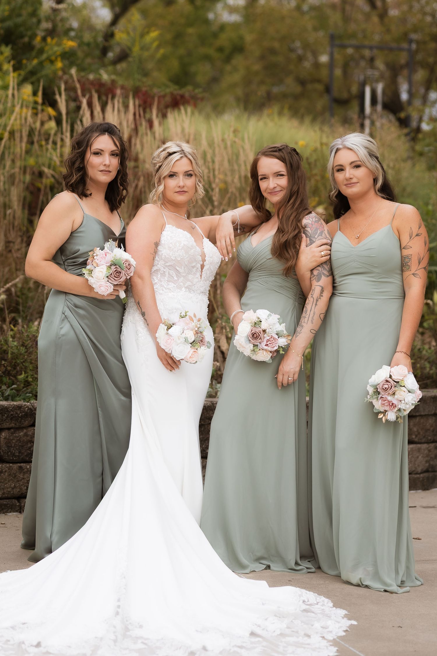 Bridesmaids in sage green dresses pose with bride in white lace gown outdoors.