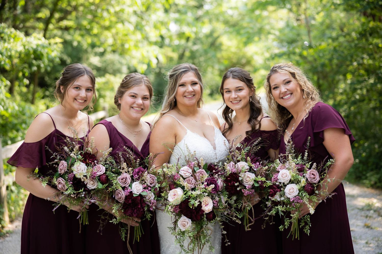 Bride in white gown with four bridesmaids in burgundy dresses holding lush purple and pink floral bouquets outdoors.