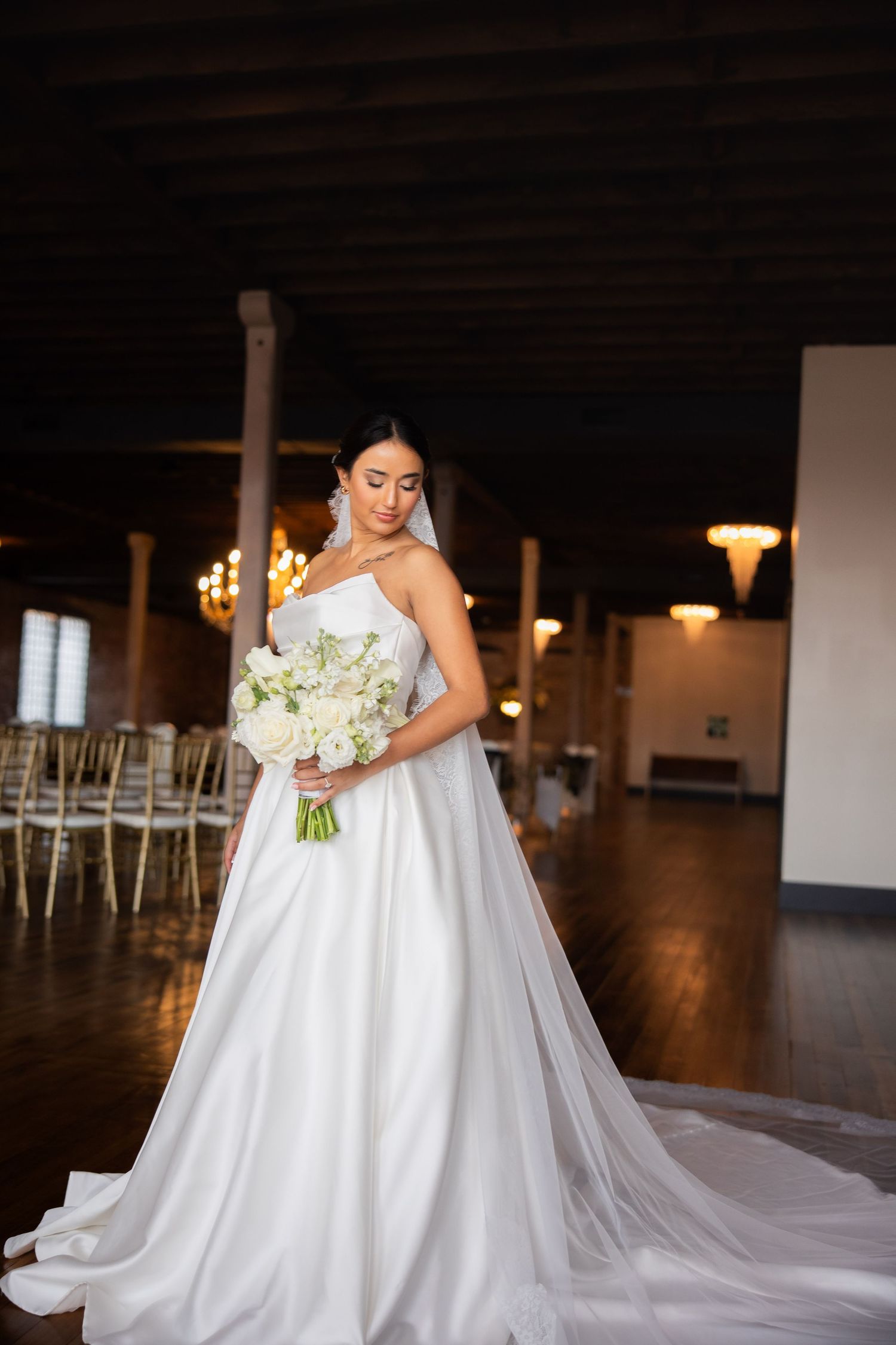 A flowing white wedding dress with a long train is displayed in an elegant indoor venue with dark wood floors.