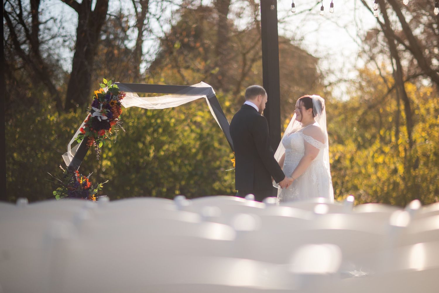 Couple exchanges vows at outdoor wedding ceremony under floral arch with white chairs and autumn foliage backdrop.