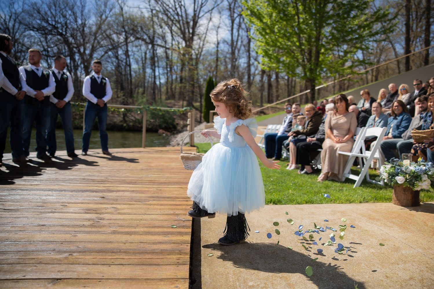 Flower girl in blue tutu dress scattering petals at outdoor wedding ceremony on wooden deck.