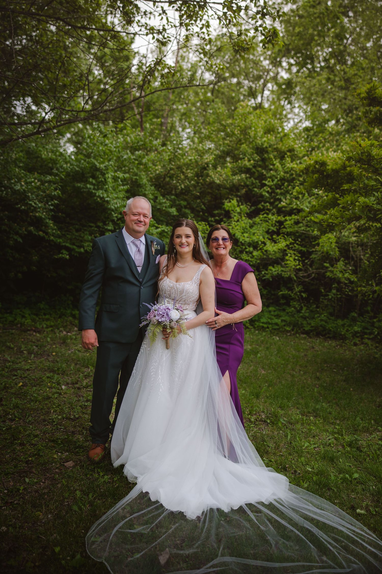 Bride in white gown poses with parents in lush green outdoor wedding setting, mother in purple dress, father in gray suit.