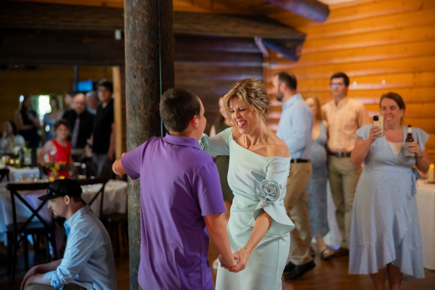Mother and son share a tender dance at a rustic log cabin wedding reception with guests watching.
