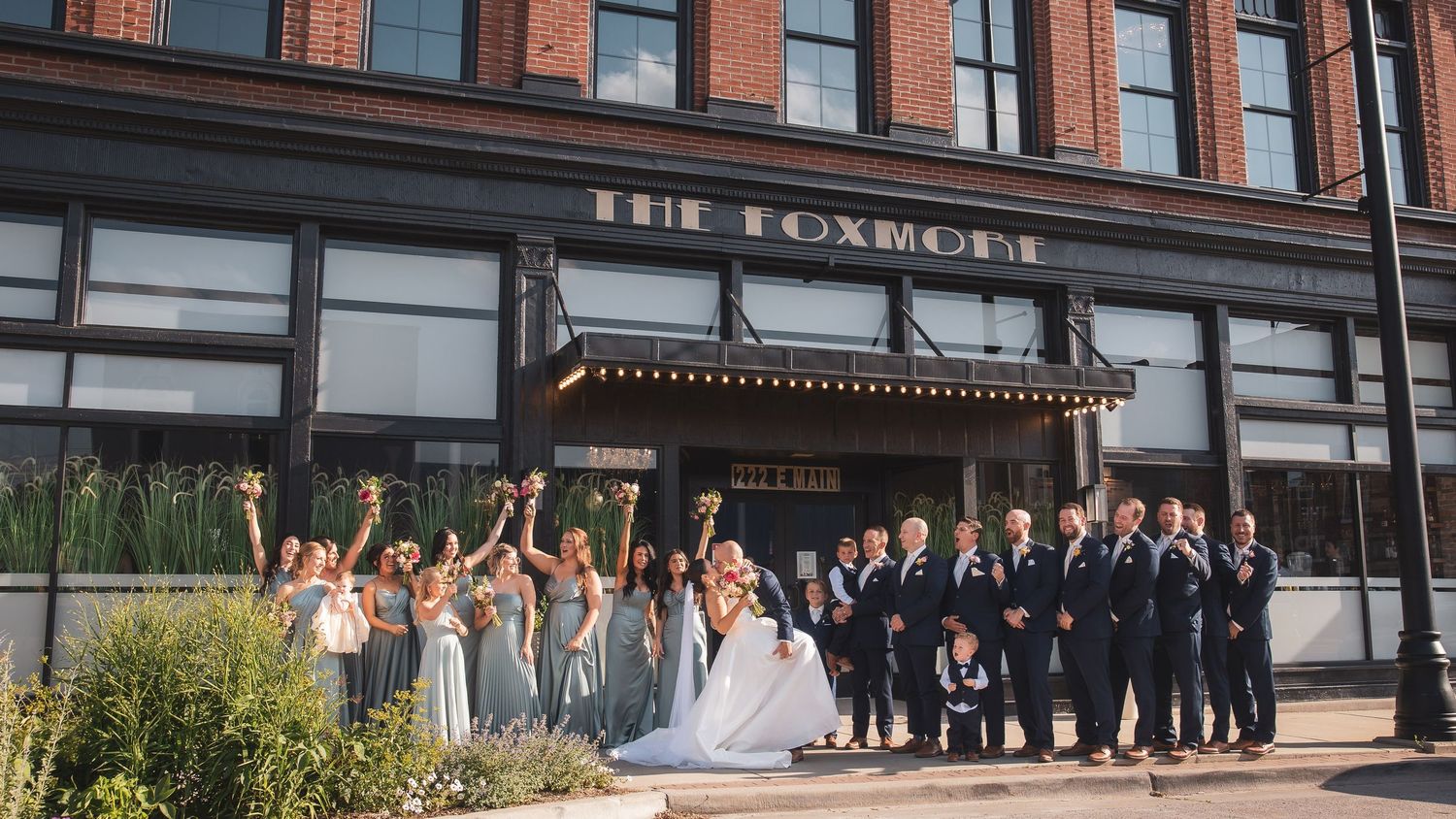 Wedding party members pose in front of a brick building on a sunny day.