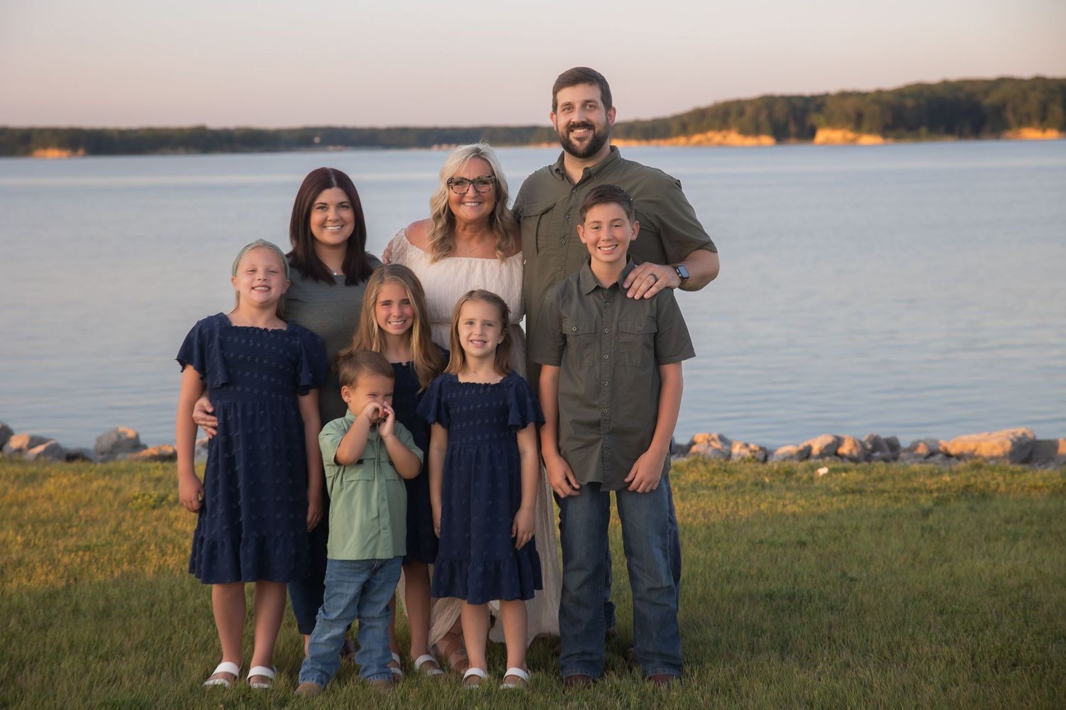 Family group poses together on grassy hill overlooking lake at sunset.