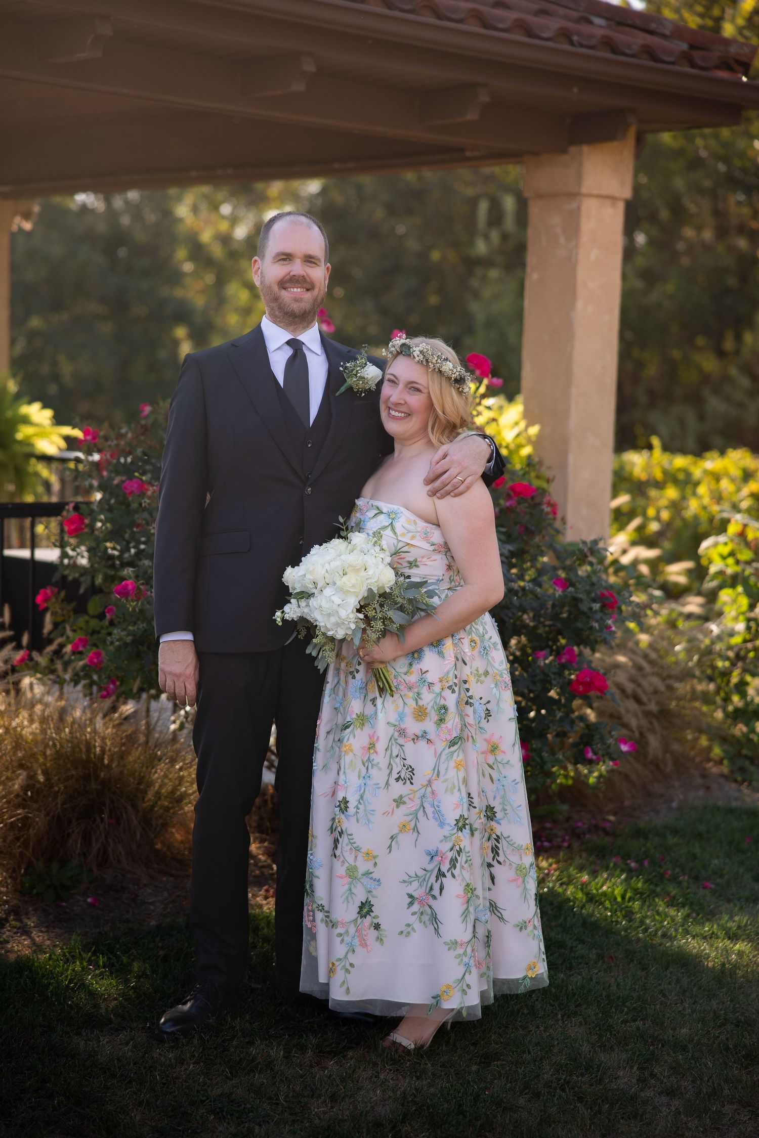 A couple poses together in a garden setting with vibrant red flowers and stone architecture.
