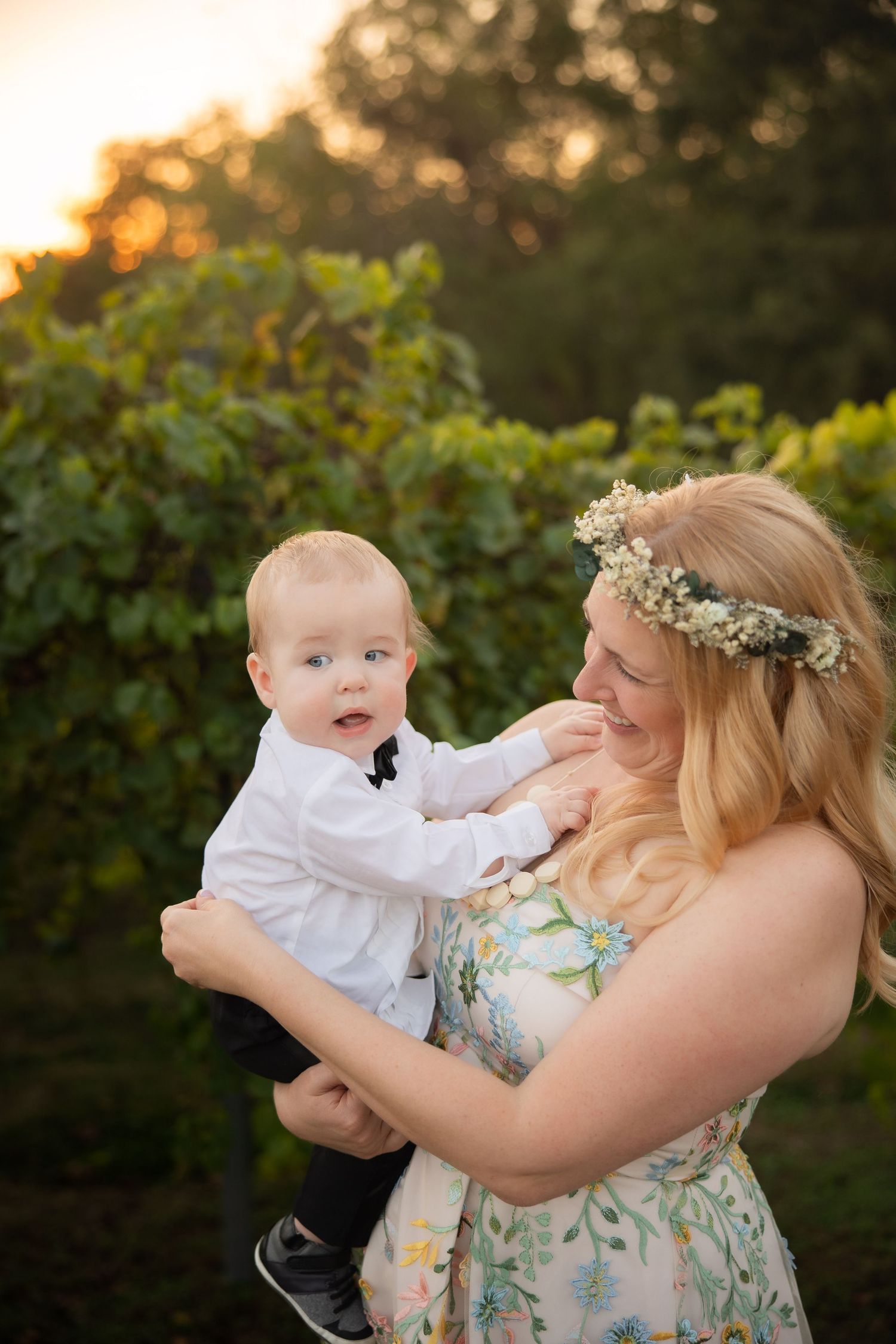 A baby in white dress shirt touches the face of someone wearing a floral dress and flower crown at sunset.