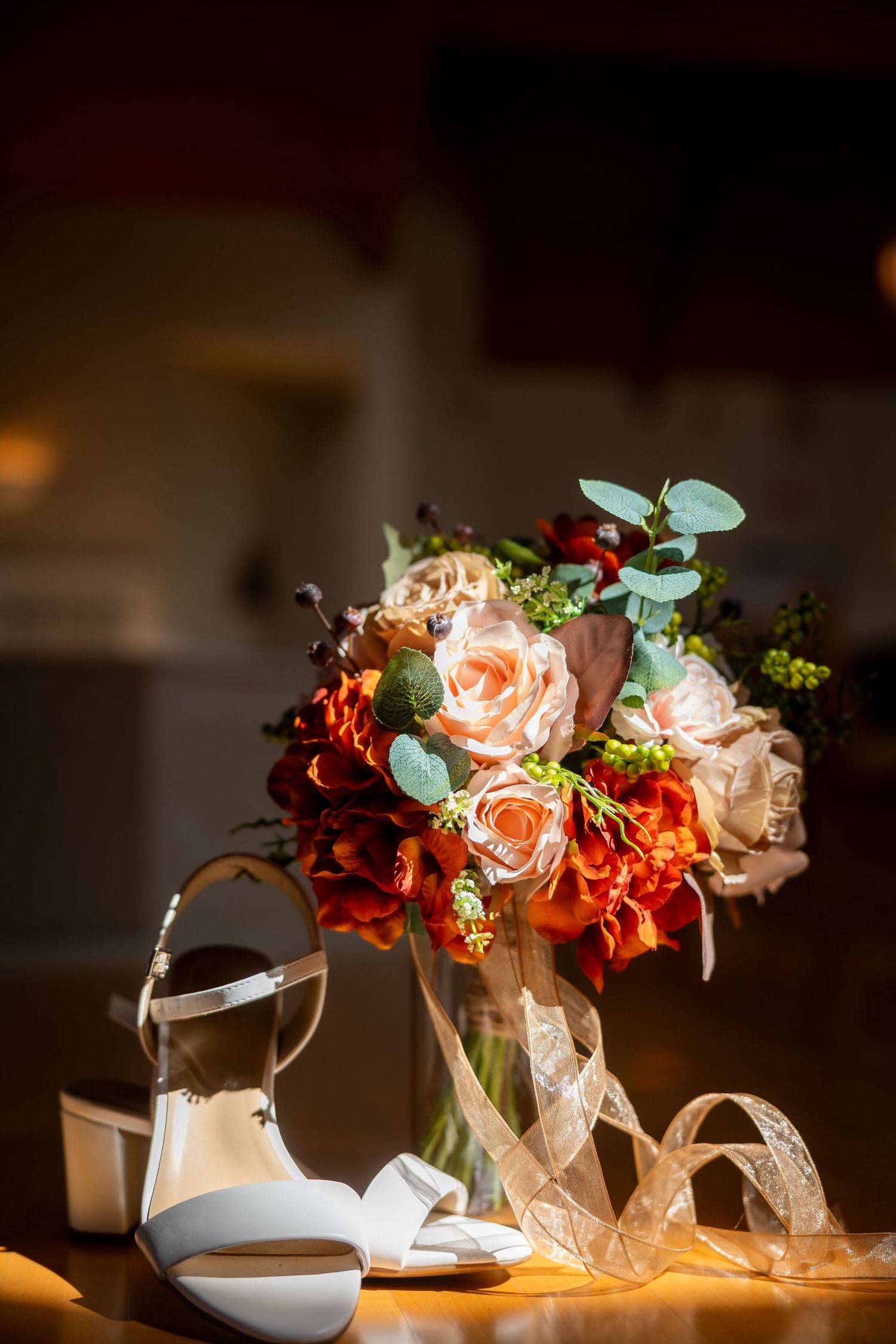 Bridal bouquet with red and peach roses beside white heeled sandals on a wooden table.