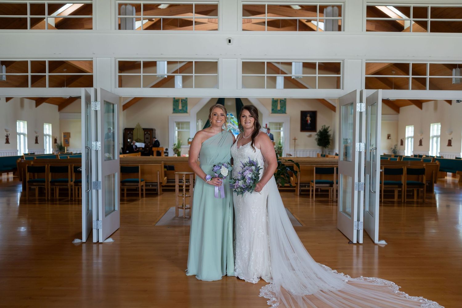 Bride in white gown and bridesmaid in sage green dress holding bouquets inside a rustic wedding venue.
