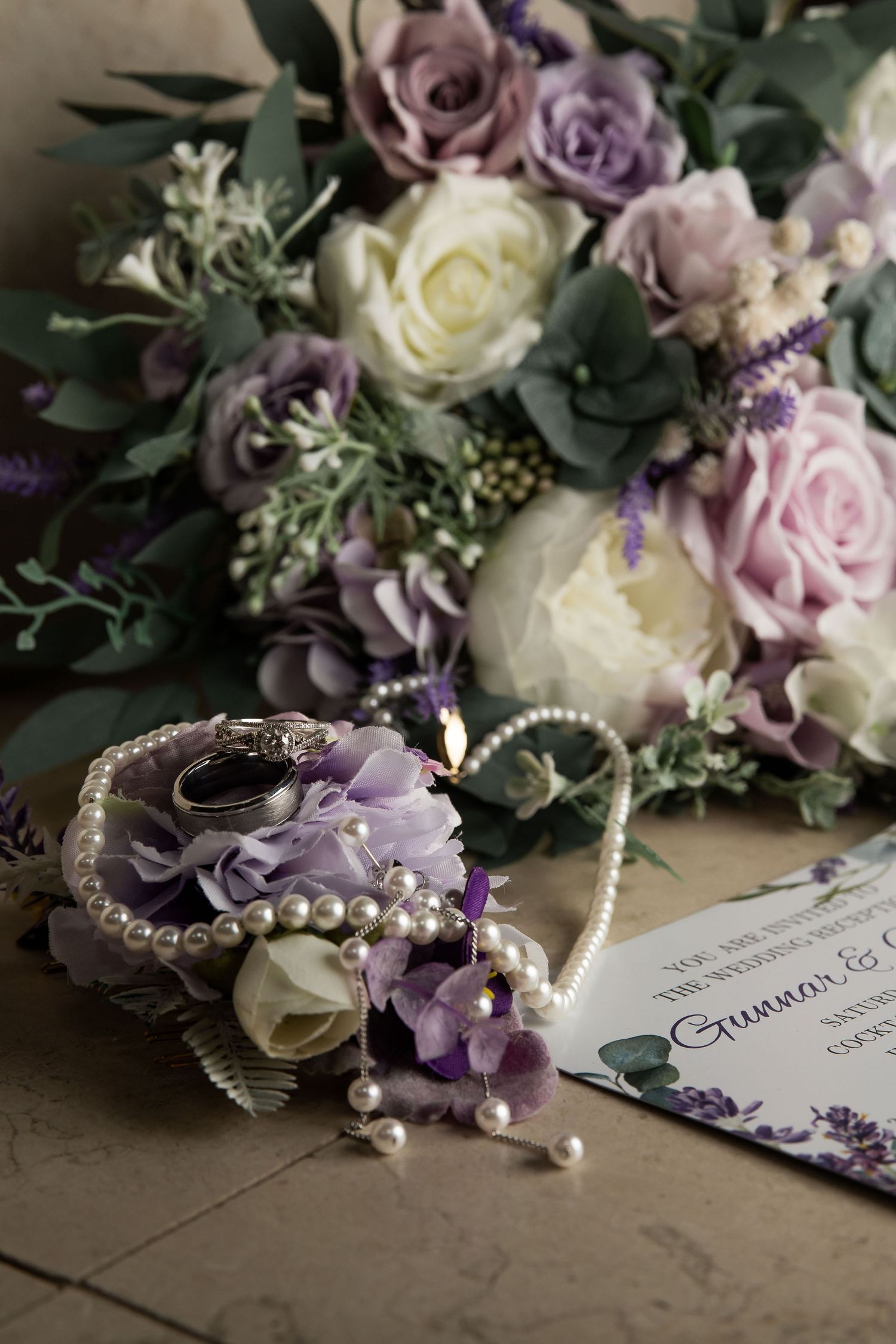 Purple and white wedding bouquet with roses, pearl jewelry, rings, and floral wrist corsage beside an elegant invitation.