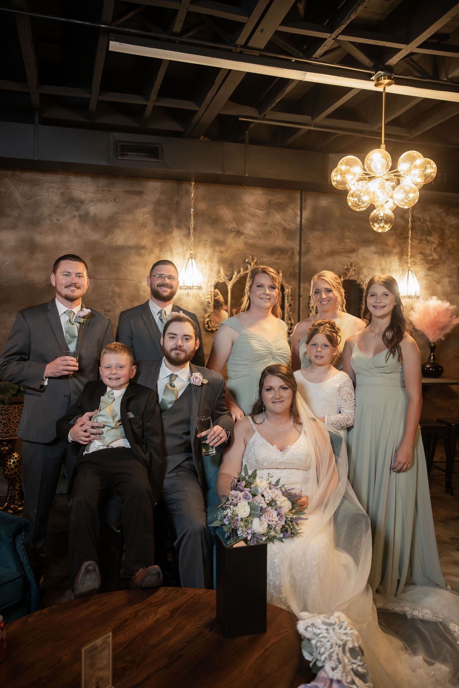 Wedding party poses together in elegant venue with chandeliers, bride holding floral bouquet surrounded by groomsmen and bridesmaids.