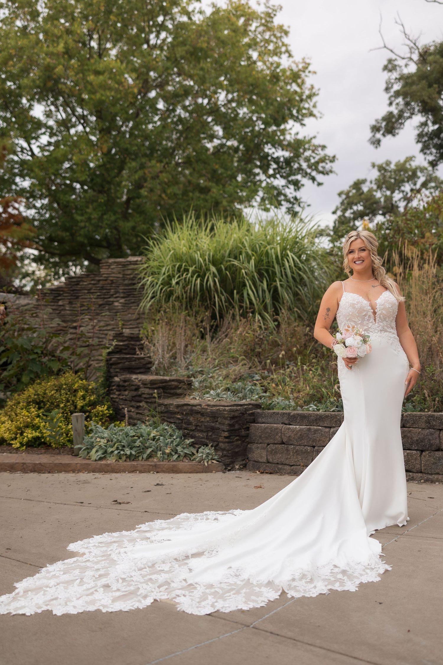 Bride in flowing lace wedding dress with long train posing against stone wall and garden landscape.
