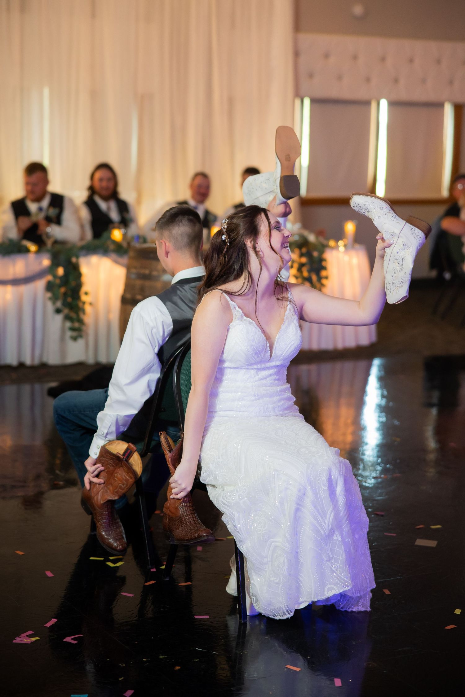 Bride and groom play the shoe game at their wedding reception, holding shoes up while guests watch and celebrate.