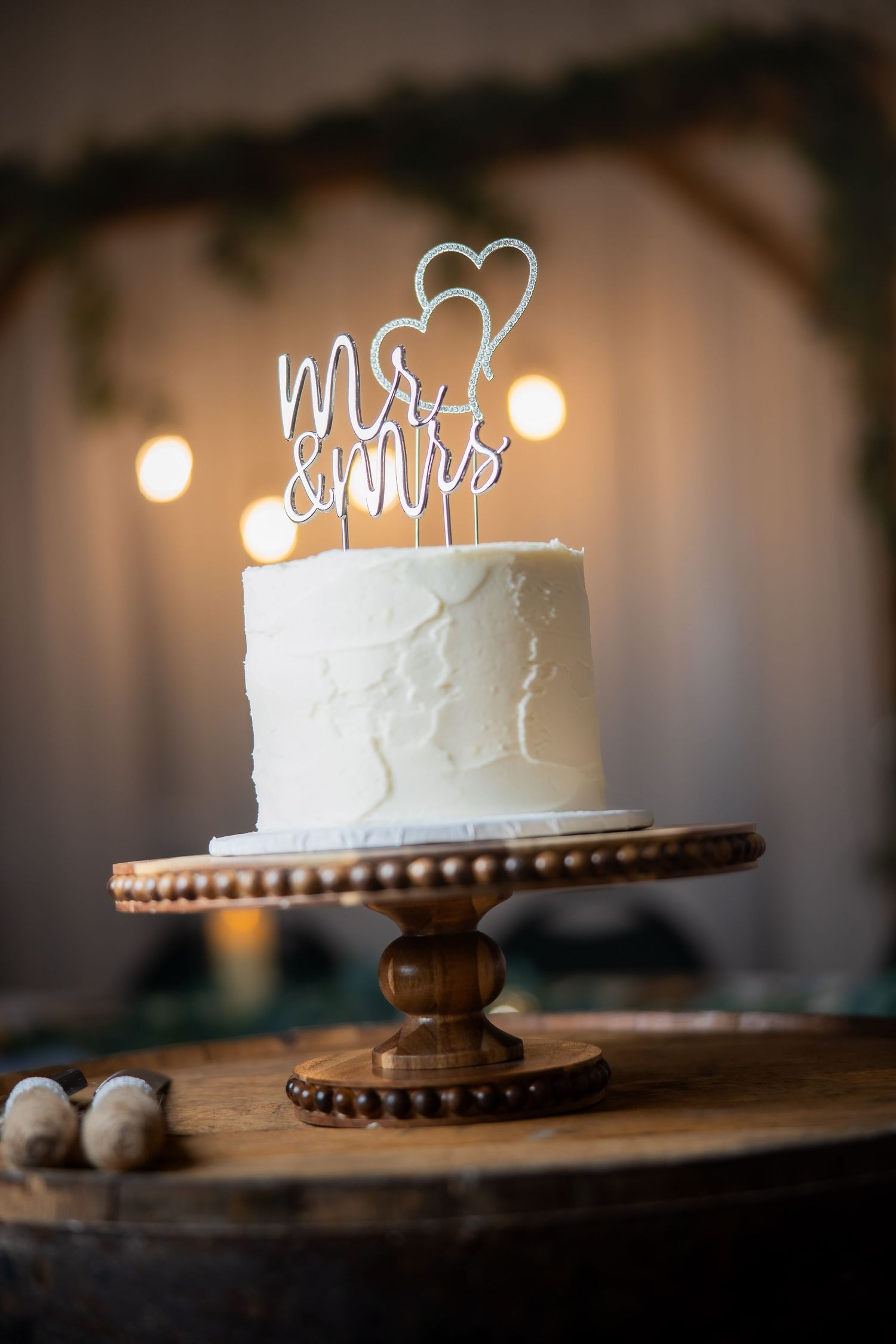 White wedding cake with Mr & Mrs topper on wooden stand, displayed against warm bokeh lights backdrop.
