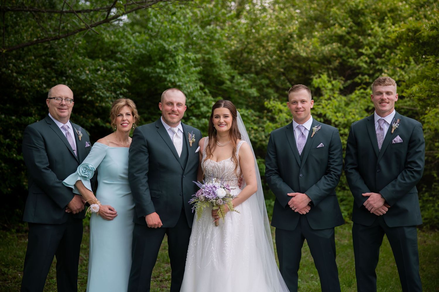 Wedding party poses outdoors in forest setting, bride holding purple bouquet with groomsmen in dark suits and family members.