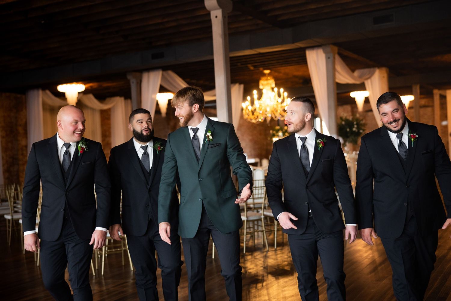 Groom in green suit walks with four groomsmen in black suits inside an elegant wedding venue with chandeliers.
