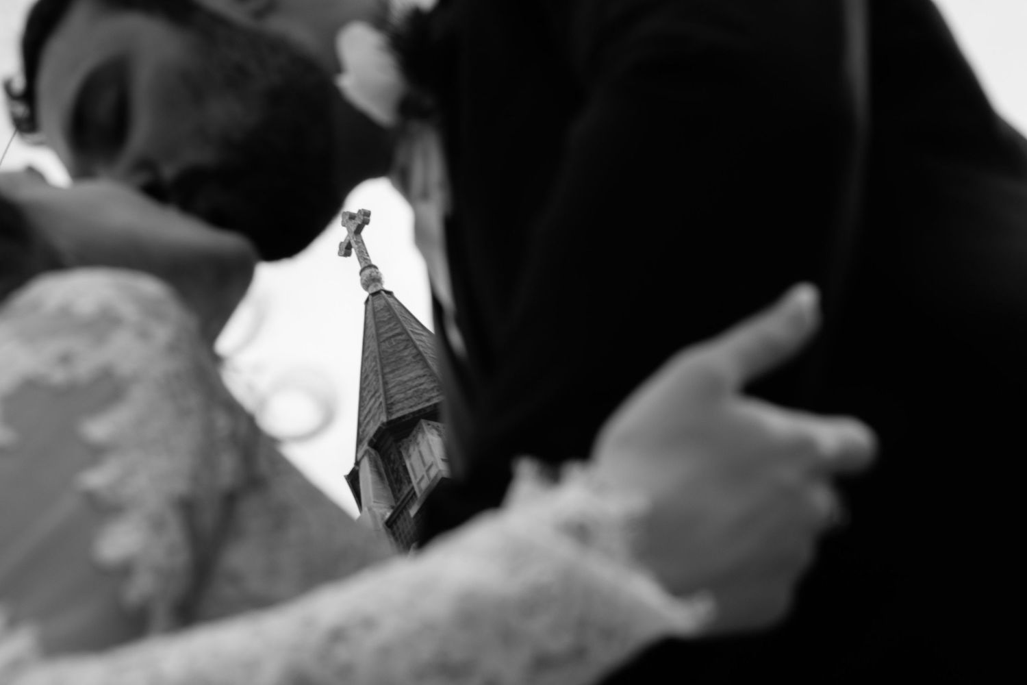 Black and white photo of bride and groom embracing with church steeple visible between them.