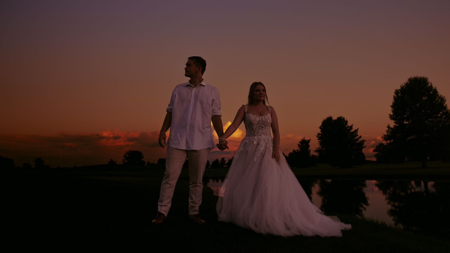 Bride and groom holding hands silhouetted against a stunning orange and purple sunset sky near a reflective pond.