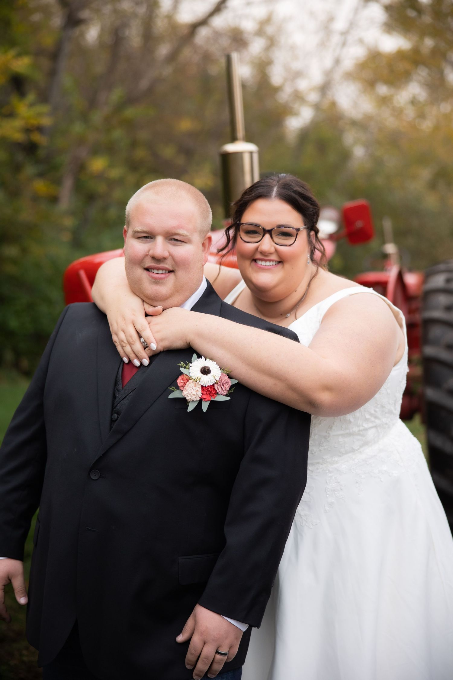 A happy couple in wedding attire poses outdoors with a red tractor visible in the background.