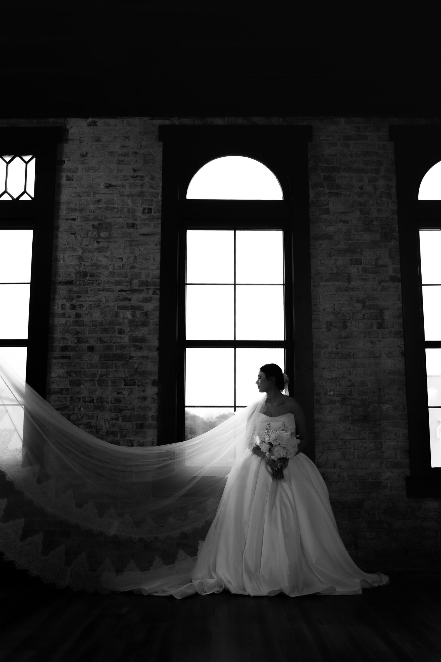 A silhouette of a bride in a flowing gown against arched windows in dramatic lighting.