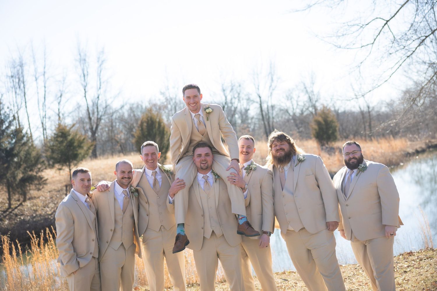 Groomsmen in tan suits pose outdoors near a pond, lifting the groom on their shoulders in a fun wedding photo.