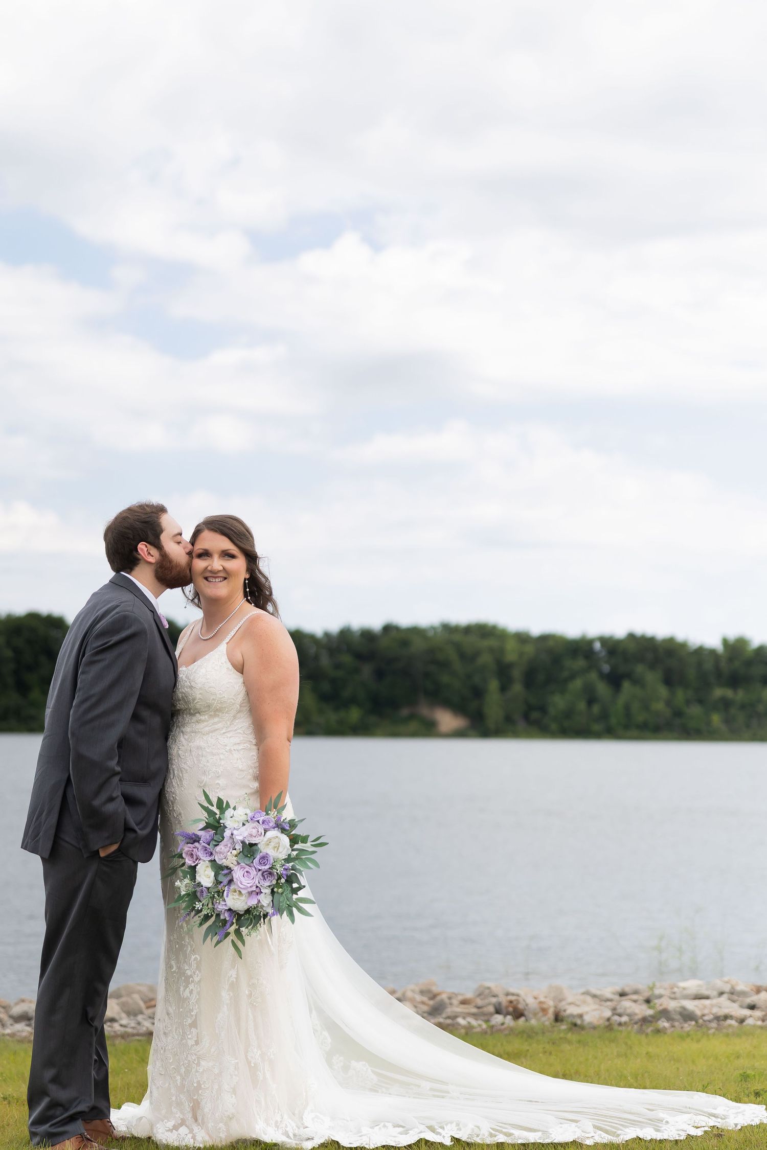 Bride in lace gown holds purple bouquet as groom kisses her cheek by a lakeside on their wedding day.