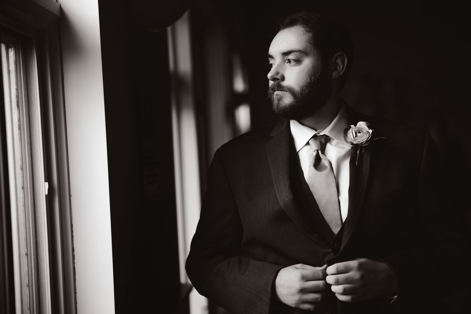 Handsome bearded groom in dark suit with boutonniere gazing out window in black and white photo.