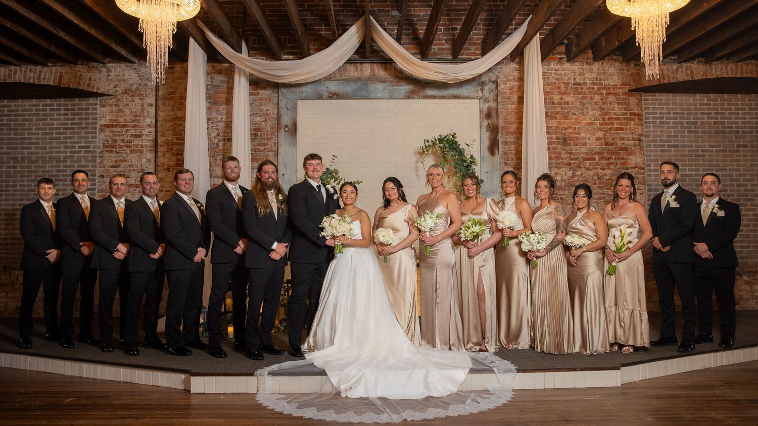 Large wedding party poses in front of draped fabric backdrop with chandeliers in rustic brick venue.