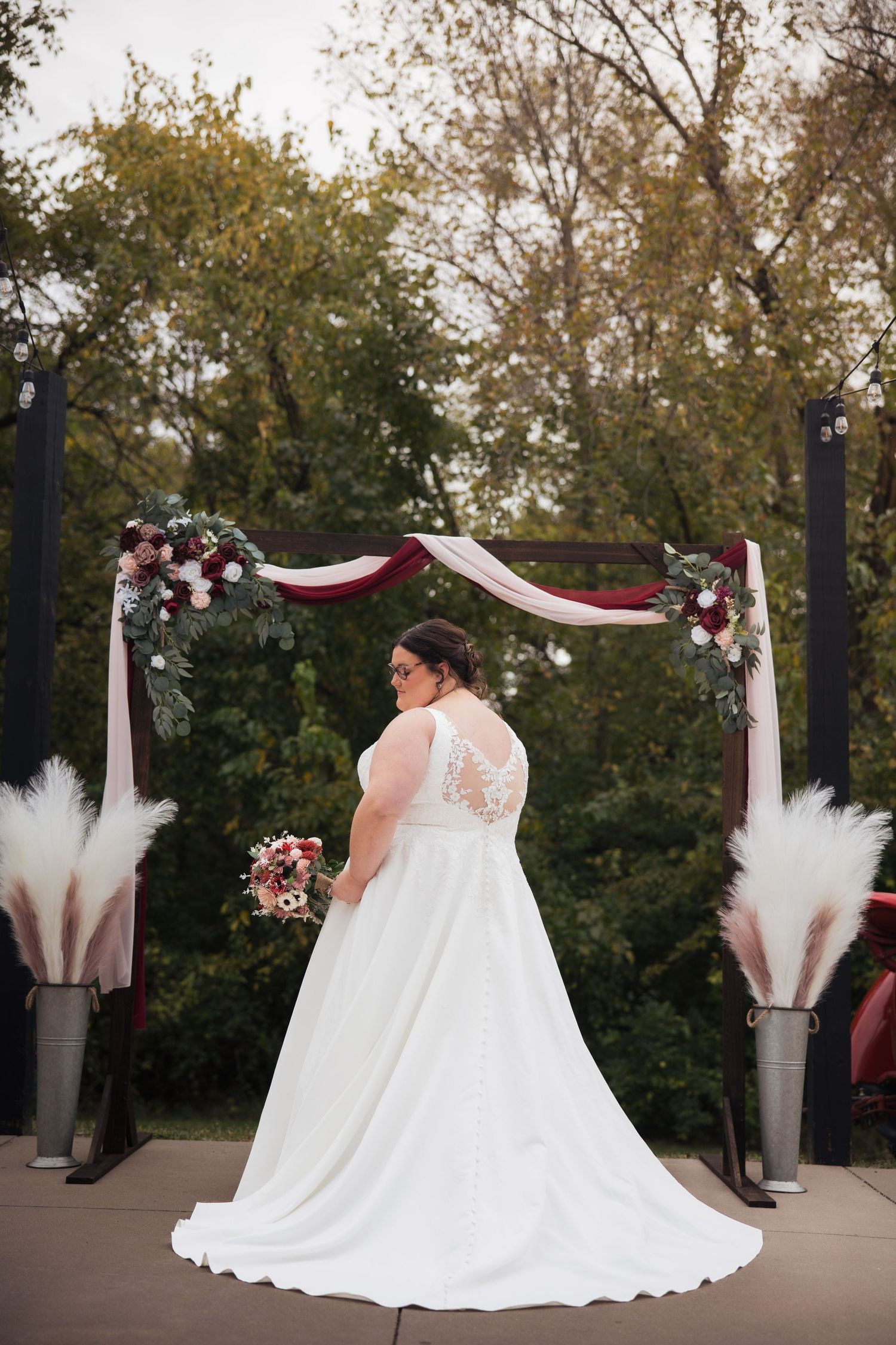 A bride in a flowing white gown poses under a decorated wedding arch with burgundy and white decor.