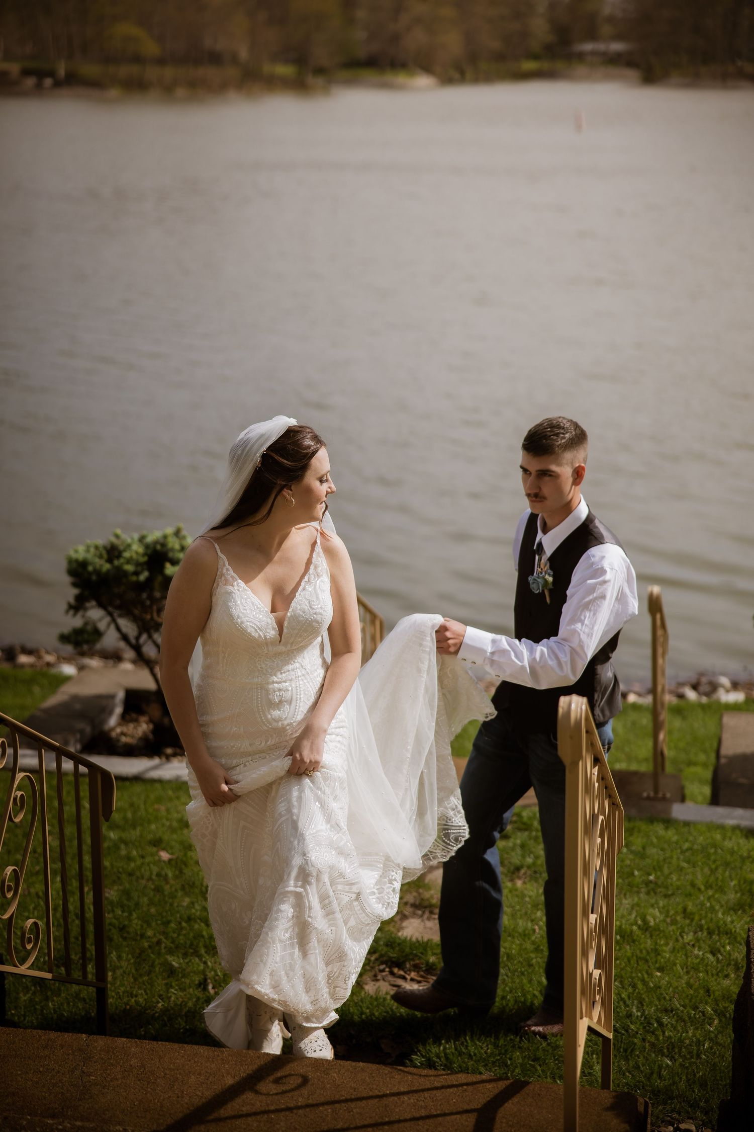 Groom carries bride's wedding dress train as couple walks up lakeside steps.